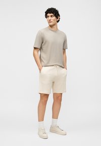 Άνδρας φοράει μπεζ μπλουζάκι, κρεμ σορτς, λευκές κάλτσες και μπεζ αθλητικά παπούτσια, στέκεται με τα χέρια στις τσέπες σε απλό φόντο.