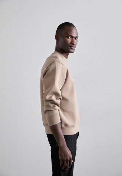 Beige genser med rund hals og ribbede mansjetter, parret med mørke jeans. Glatt stoff med subtile sømdetaljer, vist i profil.