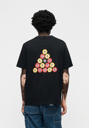 Άτομο με σκουρόχρωμα σγουρά μαλλιά και λεύκη, φοράει μαύρο μπλουζάκι με πολύχρωμο σχέδιο τριγώνου με μπίλιες μπιλιάρδου στην πλάτη, συνδυασμένο με μπλε τζιν.