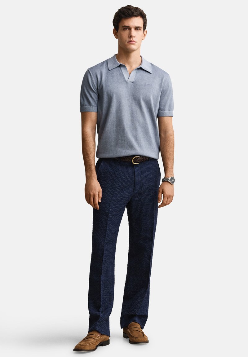 Joven con polo de manga corta azul claro, pantalones azul marino oscuros con textura, mocasines marrones, cinturón y reloj de pulsera, posando frente a un fondo blanco.