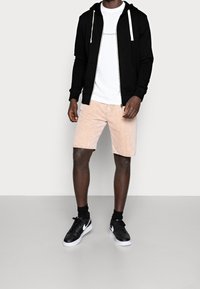 Sudadera negra con cremallera sobre una camiseta blanca, pantalones cortos de pana beige, calcetines negros y zapatillas Nike negras en una persona de pie contra un fondo blanco.