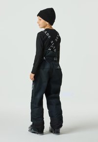 Svart skidklädsel med märkta remmar, bestående av en åtsittande långärmad topp och isolerade, löst sittande byxor. Svart mössa och stövlar.