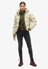 Giacca puffer beige chiaro con cappuccio, cerniera nera, polsini a costine e maglione lavorato a maglia verde. Jeans scuri e stivali marroni con lacci.