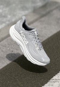 HOKA grå løpetirsko med mesh-overdel, hvit merkevare, polstret såle og teksturerte detaljer, vist i en dynamisk, hevet posisjon.