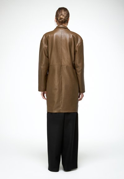 Osoba z włosami związanymi w niski kok ubrana w długi brązowy skórzany płaszcz i szerokie czarne spodnie, stojąca tyłem na jednolitym białym tle.