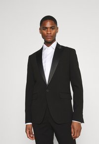 Jachetă de tuxedo neagră cu rever din satin, două buzunare frontale, închidere cu un singur nasture, croială ajustată, purtată cu o cămașă albă.