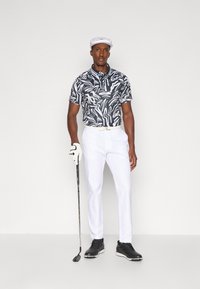 Camisa polo de manga curta com padrão preto e branco, calças brancas, boné branco, sapatos de golfe pretos, segurando um taco de golfe, com uma luva de golfe na mão esquerda.