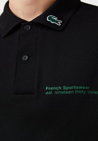 Polo noir en tissu texturé, avec un texte vert brodé "French Sportswear est. nineteen thirty three" et un logo de crocodile sur le col.