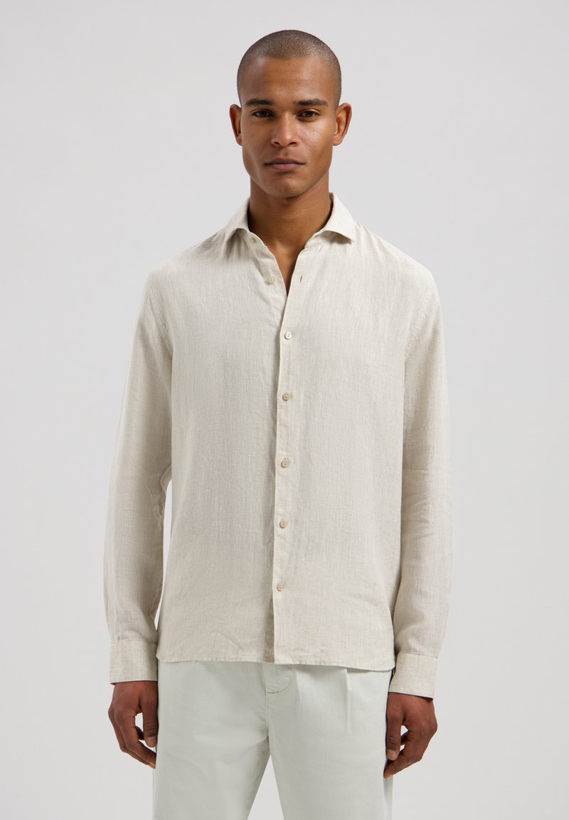 Homme aux cheveux courts portant une chemise beige à manches longues boutonnée et un pantalon clair, debout devant un fond uni.