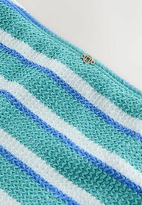 Szydełkowany koc z naprzemiennymi pasami w kolorach morskim, jasnoniebieskim i białym, o teksturowanym, falistym wzorze oraz małym metalowym akcentem.