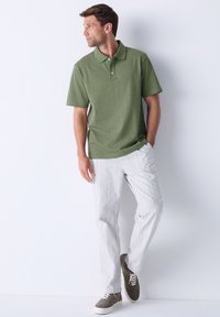 Zeleni polo s teksturiranim završetkom, uparen sa svijetlim prugastim hlačama i sivim antilop tenisicama, na bijeloj pozadini.