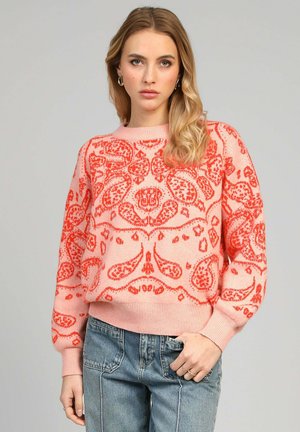 Pull rose avec un motif paisley rouge complexe, poignets et ourlet côtelés, porté avec un jean bleu clair. Tissu tricoté texturé et décontracté.