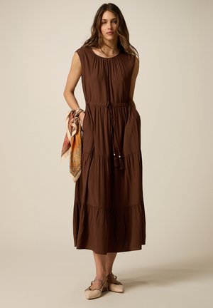 Femme portant une robe midi marron sans manches avec une taille à cordon et tenant une écharpe à motifs dans une main.