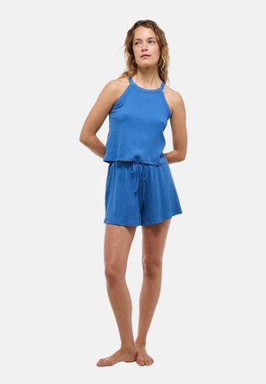 Femme debout, pieds nus, portant un haut bleu sans manches et un short bleu assorti avec un cordon, sur un fond clair uni.