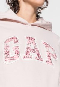 Sudadera con capucha de color rosa claro que presenta un lettering llamativo de "GAP" con un patrón geométrico rojo. Tejido suave con un diseño clásico de bolsillo canguro.