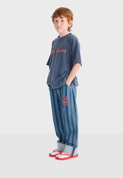 Bambino dai capelli rossi in piedi con le mani nelle tasche, indossa una maglietta grafica blu oversize, jeans a gamba larga a righe e calzini bianchi con sandali rossi.