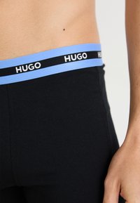Bañador negro con una cinturilla elástica de color azul claro que presenta el logotipo "HUGO" en negro repetido. Textura suave, diseño ajustado.