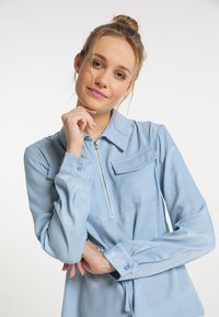 Vaaleansiniäinen vetoketjullinen pusero, jossa on pitkät hihat, kaksi rintataskua ja sileä, hieman kiiltävä pinta. Muotoiltu istuvuus hienovaraisella suunnittelulla.