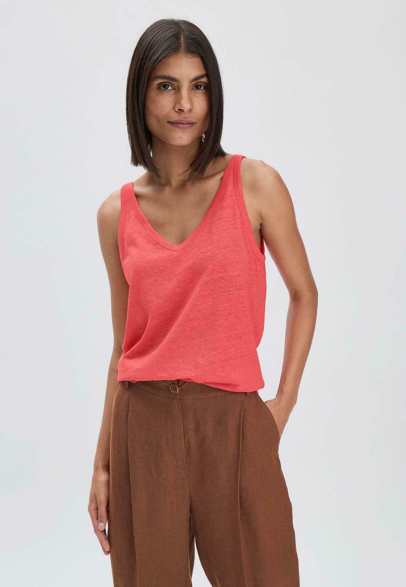 Koraalkleurige tanktop met een V-hals ontwerp, gemaakt van zacht materiaal. Gecombineerd met bruine, high-waisted broek met twee knopen en plooien.