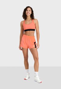Sportovní outfit se zářivě neonově oranžovým sportovním podprsenkou s černým pruhem a odpovídajícími šortkami. Lehká tkanina a elastický pas.