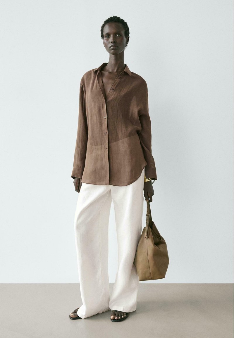 Frau trägt ein lockeres braunes Hemd mit Knöpfen, weite weiße Hosen, schwarze Sandalen und hält eine braune Handtasche vor einem schlichten hellen Hintergrund.