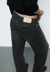 Mustat farkut looser-mallilla, joissa on nahkalaminaatti, jossa lukee "D91" takana, näkyvät ompeleet ja kaksi takataskua.