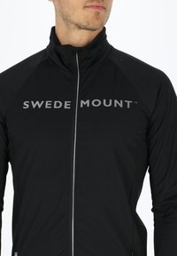 Svart idrottsjacka med hög krage, med full dragkedja, reflekterande "SWEDEN MOUNT"-logotyp och kontrasterande sidopaneler.