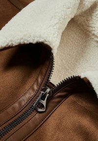 Giacca marrone con cerniera argentata parzialmente aperta, che mostra un morbido rivestimento in sherpa bianco e una rifinitura in pelle liscia attorno alla cerniera.