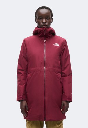 Person trägt eine lange, bordeauxrote Jacke mit Reißverschluss, hohem Kragen und Kapuze und steht vor einem schlichten, hellen Hintergrund.