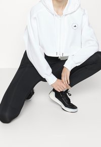 Sudadera corta blanca con cremallera frontal y detalle de logo, combinada con pantalones ajustados negros y zapatillas negras con suelas blancas.