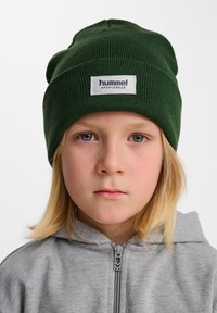 Grönstickad mössa med uppvikt kant, med en broderad vit etikett med "hummel sportswear." Mjuk textur, tät passform, avslappnad design.