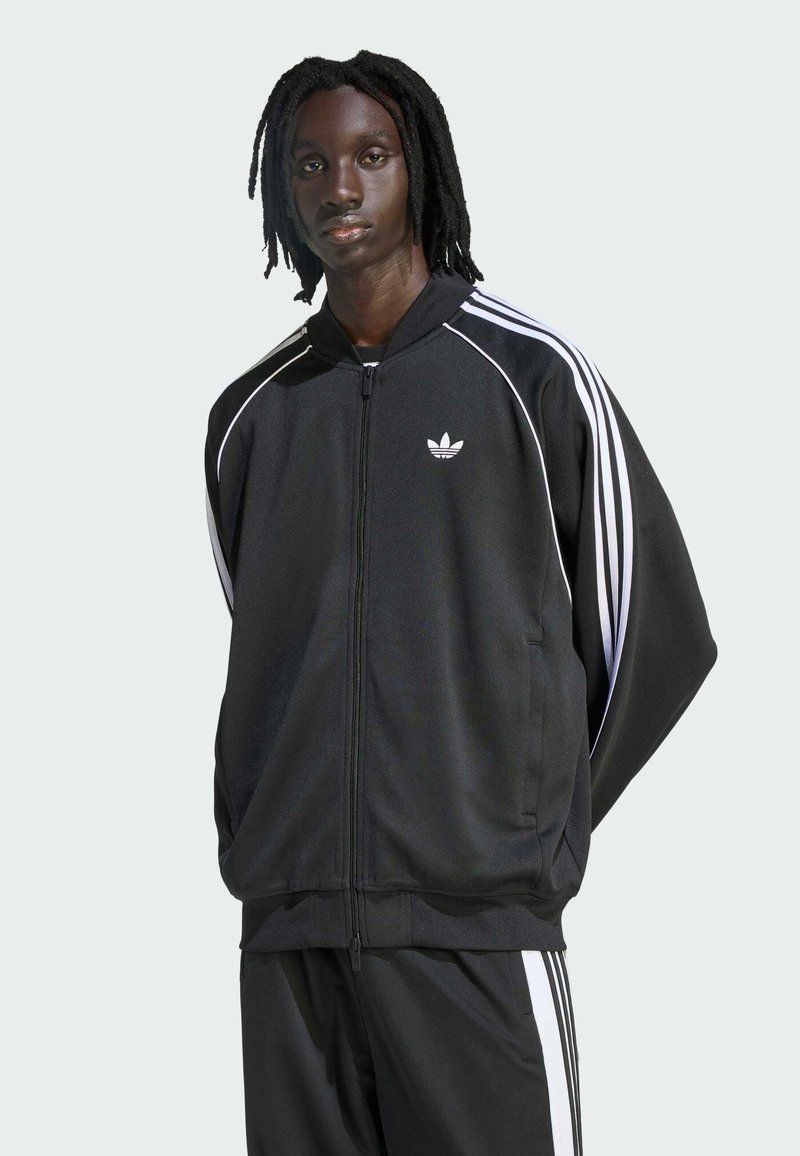 Μαύρο τζάκετ στίβου με λευκό σχέδιο τριών ριγών, με φερμουάρ μπροστά, ψηλό γιακά και μικρό λογότυπο στο στήθος. Περιλαμβάνει μαύρο παντελόνι που ταιριάζει.