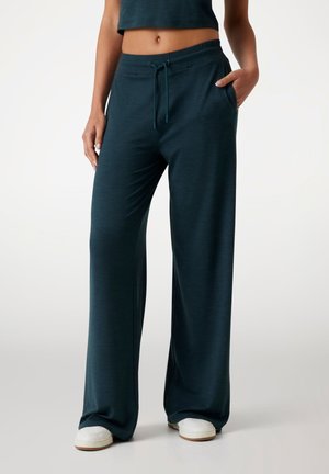 Teal wide-leg pants made of soft material with a drawstring waist, side pockets, and a small logo on the front. Casual fit. White sneakers.