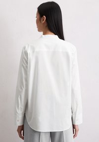 Camicia bianca a maniche lunghe con colletto, texture liscia, dettaglio con yoke sul retro e orlo arrotondato, abbinata a pantaloni grigi.