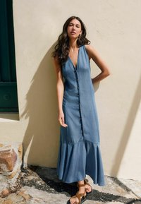 Denim maxi kjole med V-hals, knaplukning foran og udvidet kant. Har en afslappet pasform og let tekstur. Bæres med brune strapsandaler.
