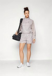 Gaiši pelēks sporta apģērbs ar rāvējslēdzēja jaku un šortiem, izgatavots no mitrumu novadoša auduma, kombinēts ar melnu duffeles somu un baltām sporta apaviem.