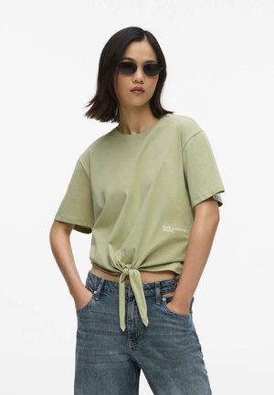 Naine, kes kannab ümmarguseid musti päikeseprille, helerohelist ees lipsuga T-särki logoga "Karl Lagerfeld Jeans" ja siniseid teksaseid, käed taskutes.