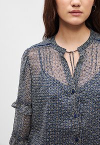 Διάφανη μπλούζα με μπλε και χρυσό γεωμετρικό σχέδιο, κουμπάκι μπροστά και ριχτές μανίκες, με λεπτομέρεια κλειδώματος στον λαιμό.