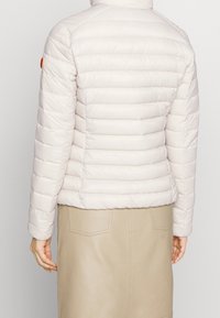 Light-colored puffer jacket with horizontal quilting, high collar, and a subtle orange logo patch on the left shoulder. Soft texture.