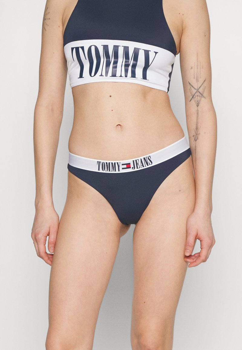 Mörkblå bikini-set med en kort topp med "TOMMY"-logotyp och matchande botten med "TOMMY JEANS"-midja. Slät tyg, sportig design.