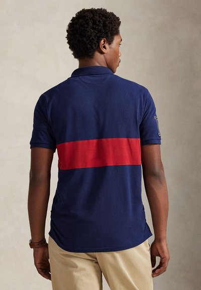 Mornarska polo majica s istaknutim crvenim horizontalnim prugom na leđima, kratkim rukavima i klasičnim ovratnikom.