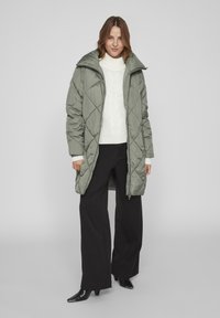 Grüner gepolsterter Mantel mit Rautensteppung, hohem Kragen und Reißverschluss vorne, getragen über einem weißen Strickpullover und schwarzen weiten Hosen.