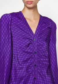 Lila blus med en strukturerad hexagonal mönstring, djup V-ringning, rynkade detaljer och knäppningar längs framsidan.