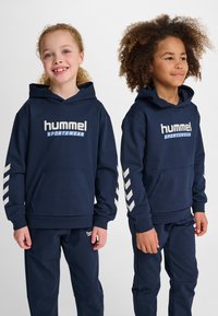Felpe blu navy con cappuccio e tasche a marsupio, caratterizzate dal logo bianco "hummel SPORTSWEAR" e dal motivo a chevron sulle maniche, abbinate a pantaloni coordinati.