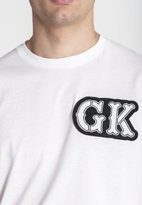 Biała bawełniana koszulka z dużą czarną haftowaną naszywką z napisem "GK" w pogrubionej, stylizowanej czcionce po lewej stronie klatki piersiowej.