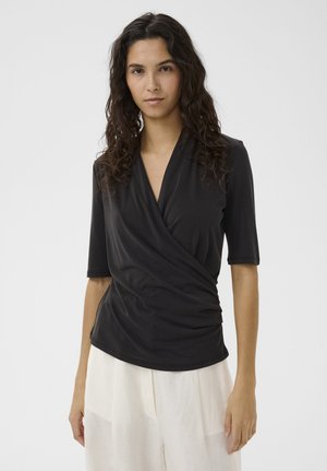 Pikakarvaline naine kannab musta mähkelaadset toppi küünarvarre pikkuste varrukatega ja valgeid laia säärega pükse, seistes ühevärvilise tausta ees.