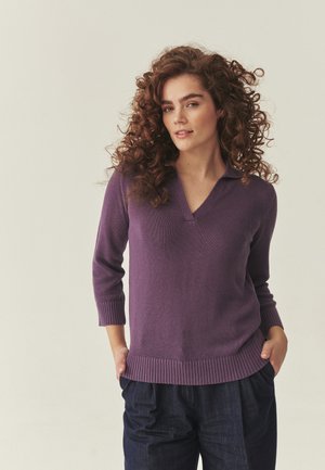 Ženska s kodrastimi lasmi, oblečena v vijoličen pulover z V-izrezom in temne hlače, stoji z rokami v žepih na enostavni podlagi.