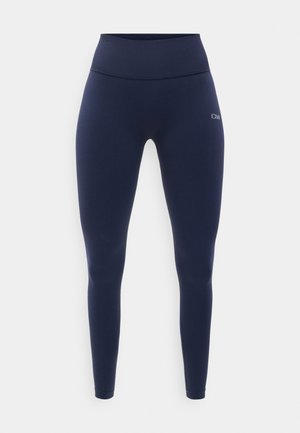 Tengerészkék, deréktól magasabb szabású, teljes hosszúságú leggings varrás nélküli kialakítással és apró fehér logóval a jobb csípőn, fehér háttér előtt.