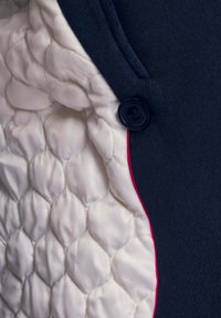 Interno di una giacca blu navy con una morbida fodera bianca trapuntata a motivo a diamante, rifinita da una sottile bordatura rossa e un bottone nero rotondo.