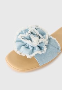 Blue denim slide sandal with a textured floral accent, frayed edges, and a tan rubber sole. Features a smooth, contoured footbed.